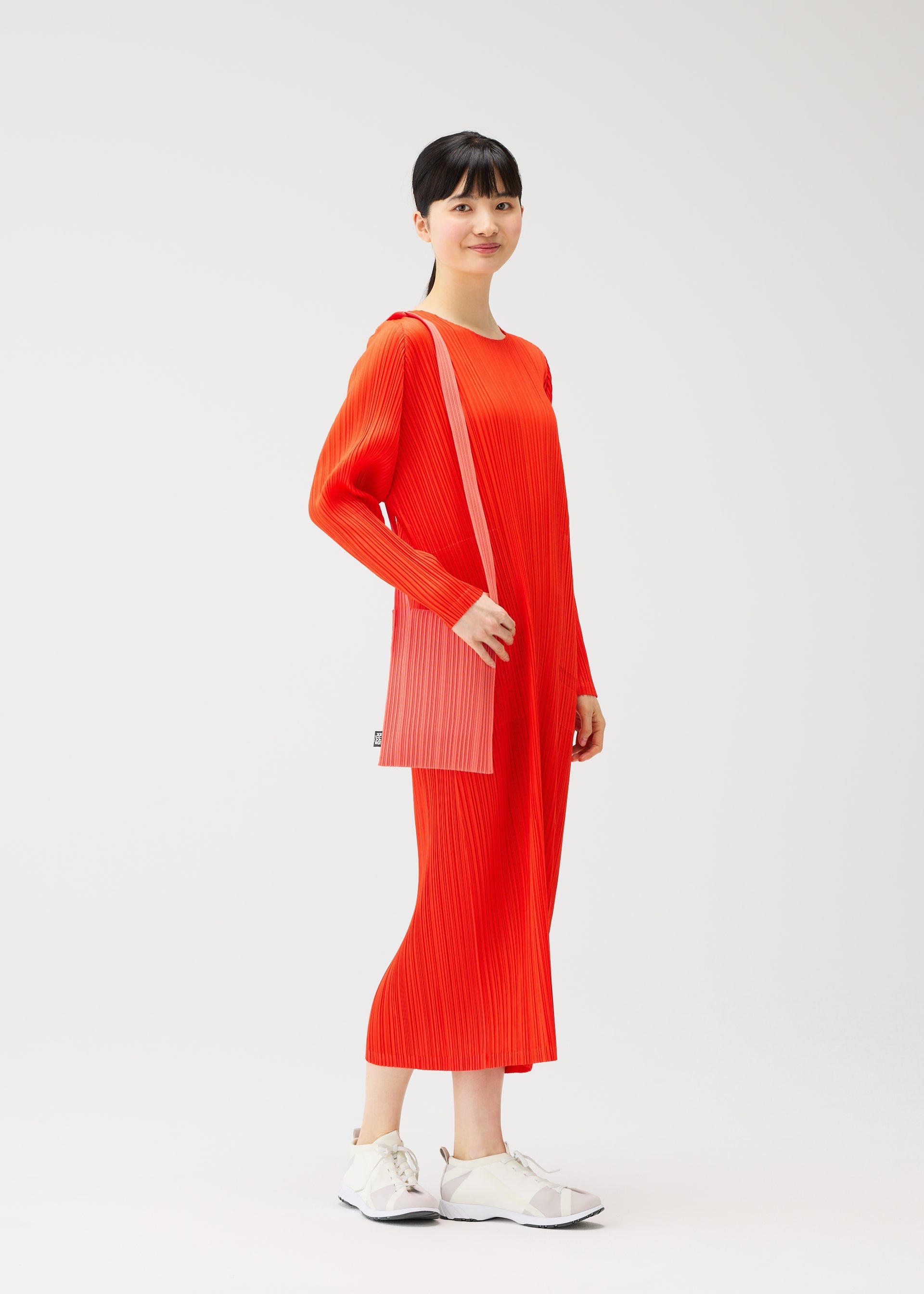 Zoomed view A woman smiles facing forward against a white background, wearing a bright red, long-sleeve pleated dress with matching mid-length skirt, white sneakers, and carrying the pink CREPE KNIT BAG by PLEATS PLEASE ISSEY MIYAKE over her shoulder.