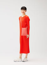 A person wears a long-sleeved bright orange dress, white sneakers, and carries the CREPE KNIT BAG by PLEATS PLEASE ISSEY MIYAKE, standing against a plain white background with a slight smile.