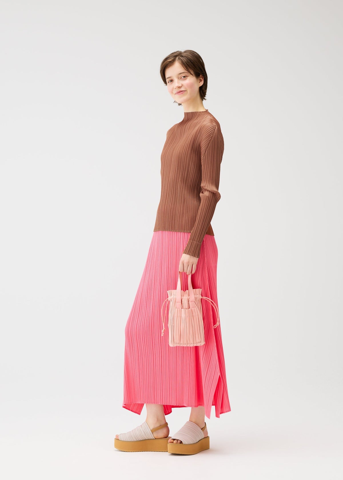 A woman stands against a plain background wearing a brown ribbed sweater, long pink pleated skirt, beige platform sandals, and holding the PLEATS PLEASE ISSEY MIYAKE PLEATS TOTE BAG. She looks at the camera with a slight smile. view 7 of 7