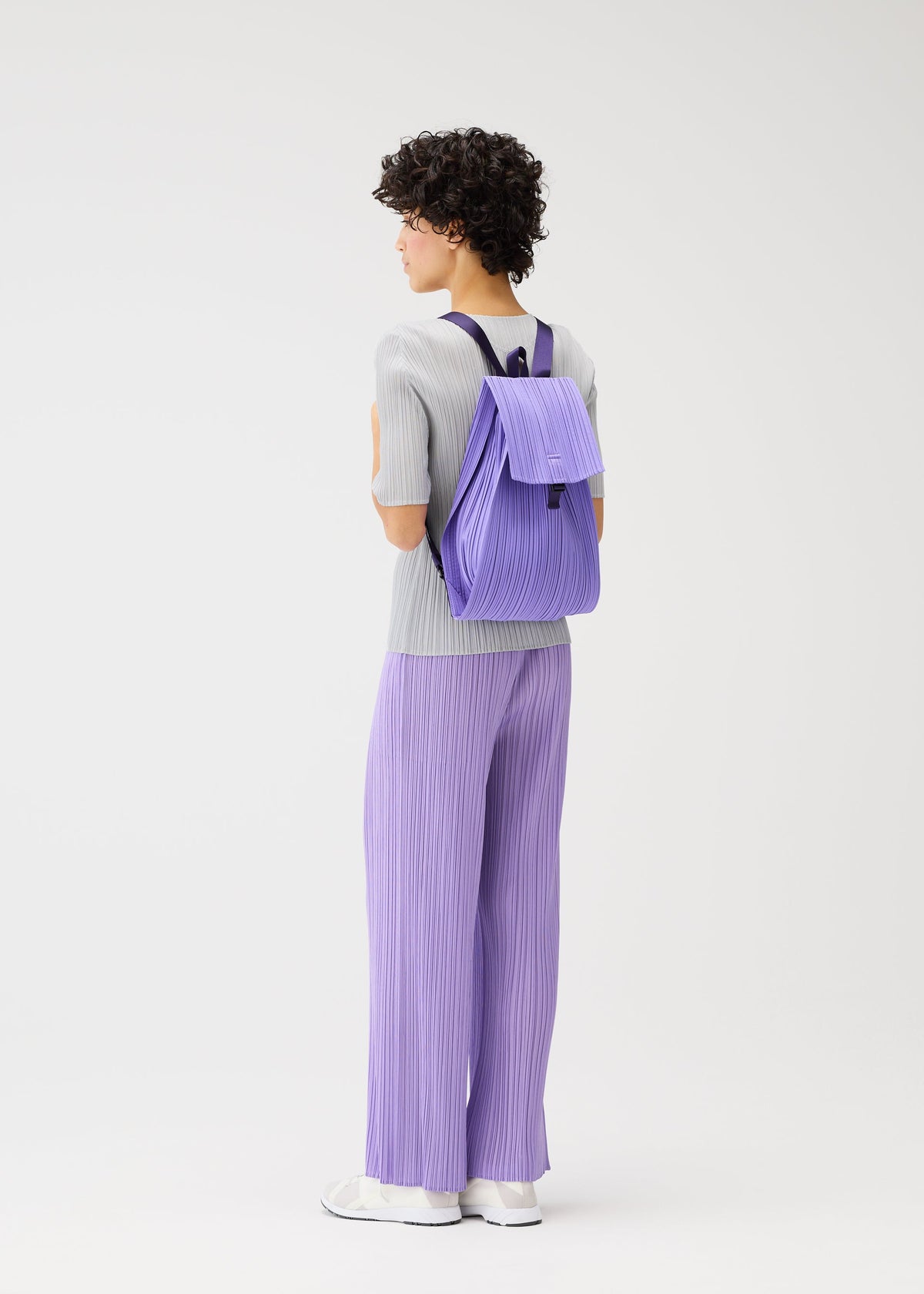 A person with short curly hair in a light grey top, lavender pleated pants, and white shoes carries the PLEATS PLEASE ISSEY MIYAKE PLEATS KNAPSACK, standing in a minimalist white studio setting facing away. view 7 of 7