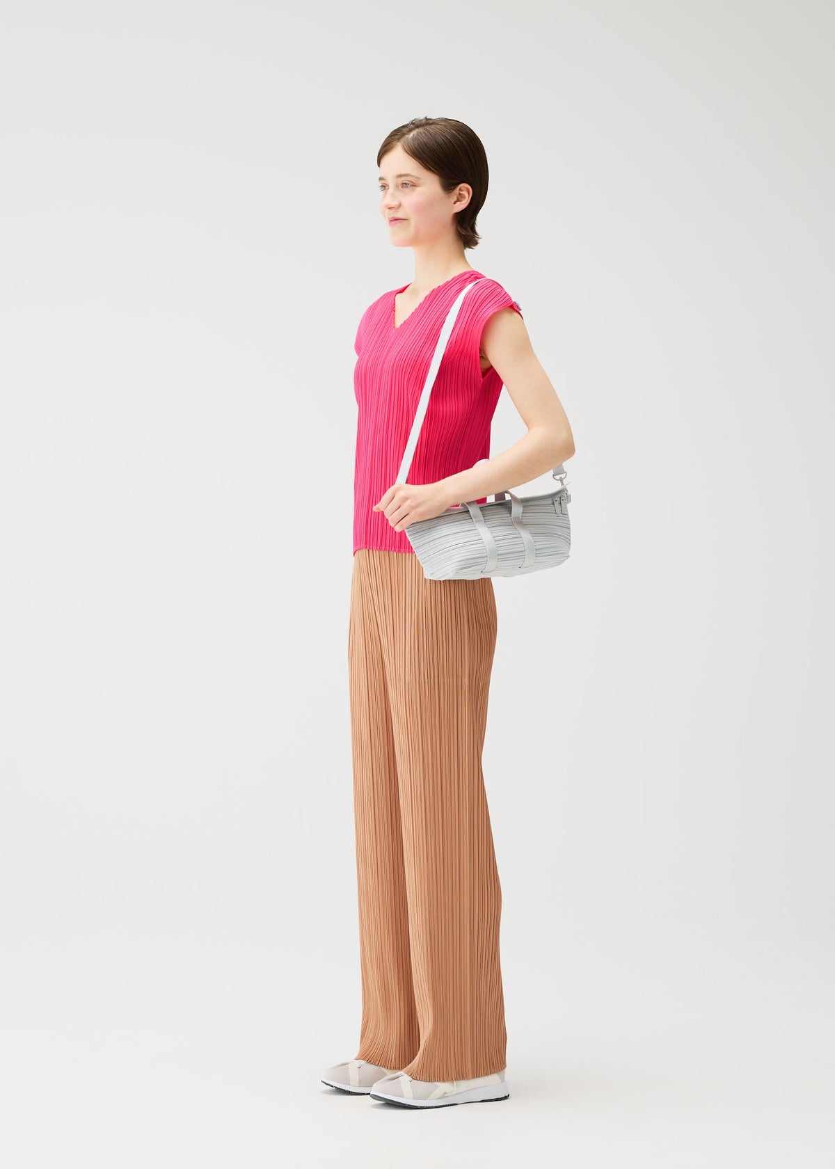 A person with short brown hair stands in a bright, minimal space wearing a pink short-sleeve top, tan pleated pants, white sneakers, and carrying the PLEATS PLEASE ISSEY MIYAKE PLEATS BOSTON BAG in light gray, smiling slightly to the side. view 7 of 8