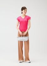 A woman stands against a plain white background, wearing a bright pink top, pleated light brown pants, white shoes, and holding the PLEATS BOSTON BAG by PLEATS PLEASE ISSEY MIYAKE with both hands.