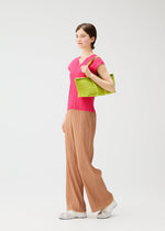A person stands against a white background, wearing a bright pink top, loose tan pleated pants, white shoes, and carrying the PLEATS BOSTON BAG by PLEATS PLEASE ISSEY MIYAKE over their shoulder.
