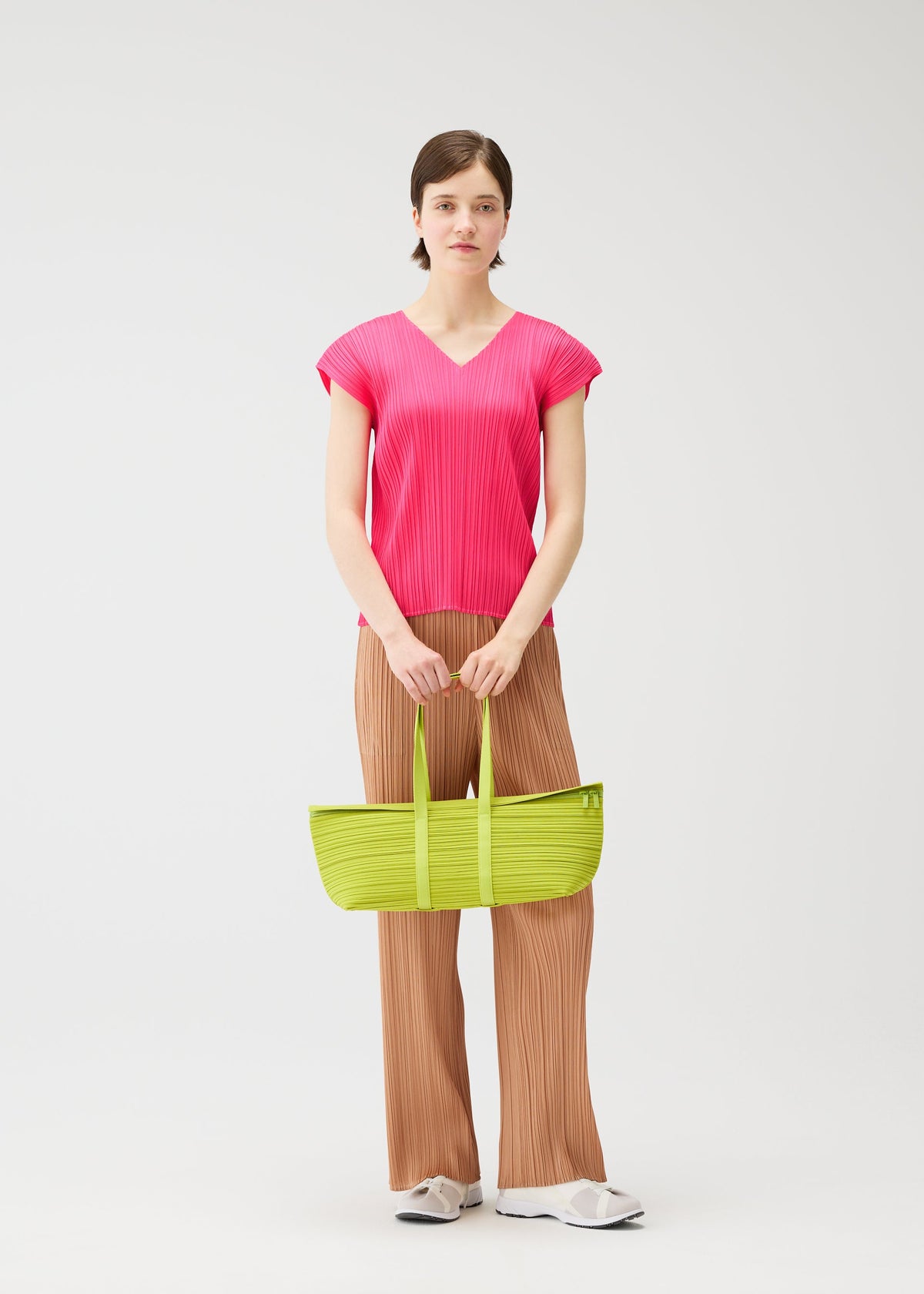 A person stands against a plain background wearing a bright pink, short-sleeve, pleated top, brown pleated pants, white shoes, and holding the PLEATS PLEASE ISSEY MIYAKE PLEATS BOSTON BAG in lime green with both hands. view 7 of 7