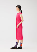 A woman with long dark hair in a ponytail stands in profile, wearing a sleeveless bright pink dress and PLEATS PLEASE ISSEY MIYAKE MARKET SOCKS, paired with black slip-on shoes against a plain white background.