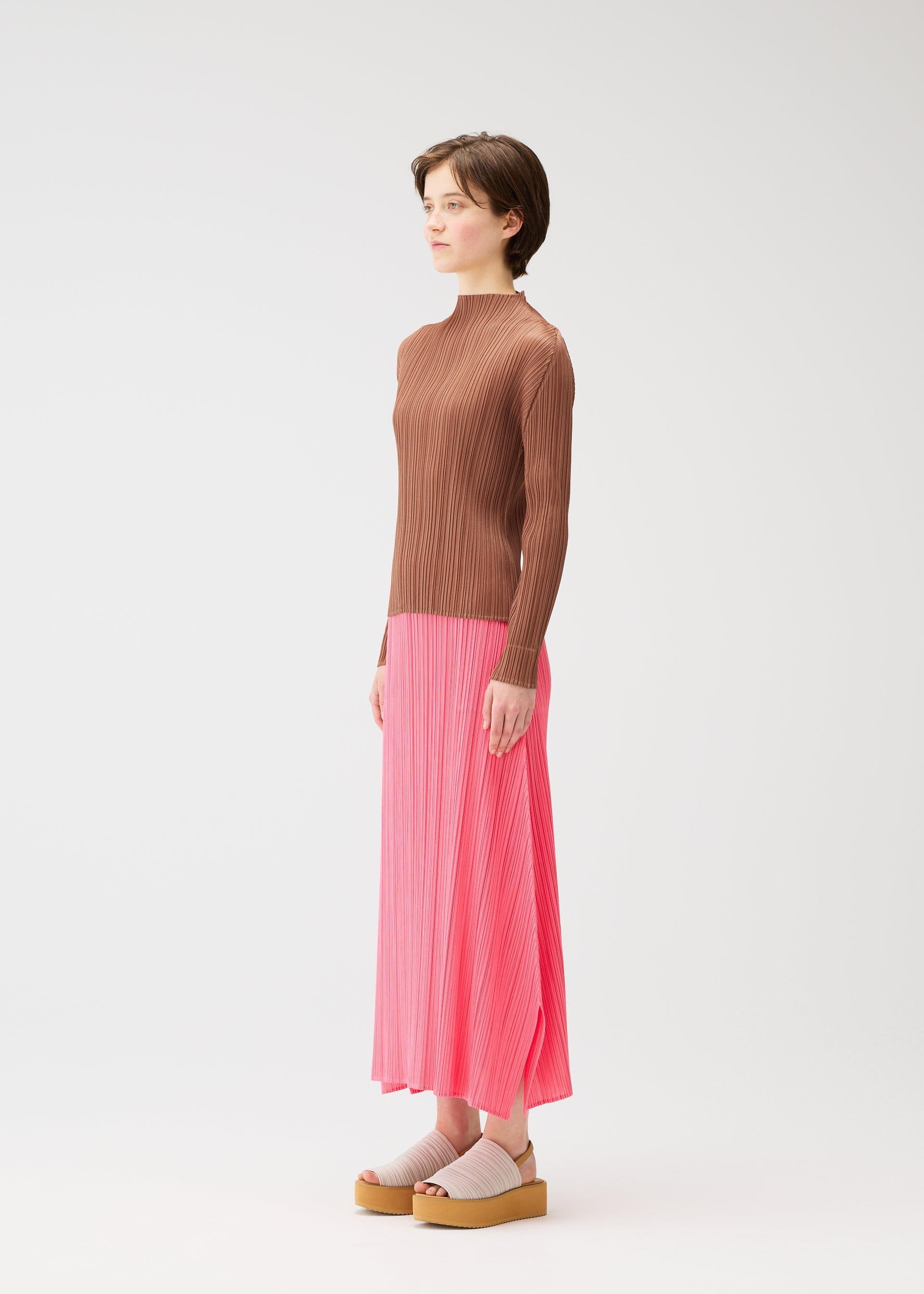 Zoomed view A person stands in profile against a plain white background, wearing a ribbed brown long-sleeve top, a pink pleated skirt, and PLEATS PLEASE ISSEY MIYAKE PLEATS KNIT SANDALS with socks.