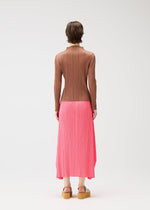 A person with short brown hair models PLEATS PLEASE ISSEY MIYAKE's PLEATS KNIT SANDALS, a ribbed brown long-sleeved top, and a pleated pink midi skirt, standing against a plain white background.
