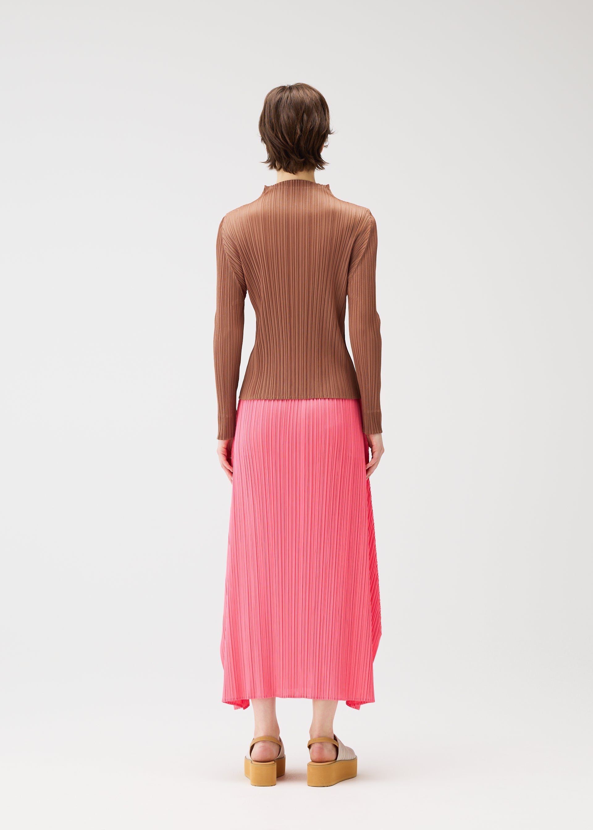 Zoomed view A person with short brown hair models PLEATS PLEASE ISSEY MIYAKE's PLEATS KNIT SANDALS, a ribbed brown long-sleeved top, and a pleated pink midi skirt, standing against a plain white background.