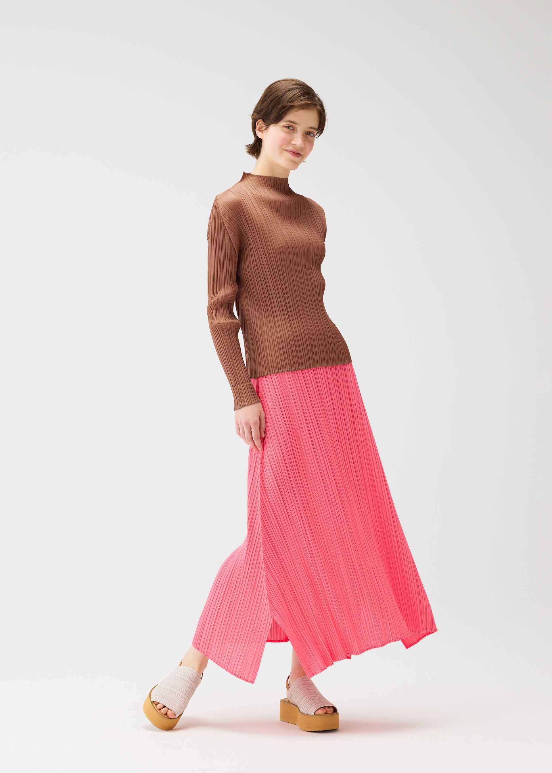 Zoomed view A person stands against a white background wearing a PLEATS PLEASE ISSEY MIYAKE long-sleeved brown knit top, a flowing pleated bright pink skirt, and thick-soled PLEATS KNIT SANDALS. They have short hair, are smiling, and stand with one foot slightly forward.