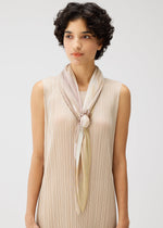 A woman with short, curly dark hair wears a sleeveless beige pleated dress and a light scarf secured with the SHELL SCARF RING by PLEATS PLEASE ISSEY MIYAKE, standing against a plain light background.