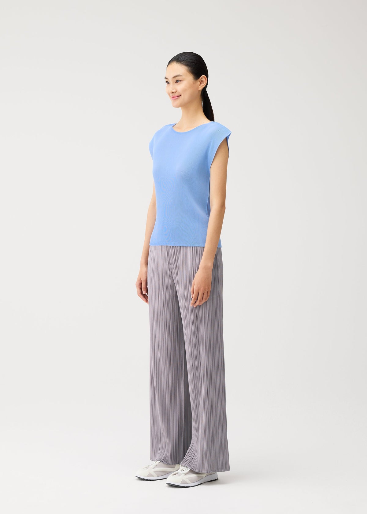 A woman stands facing left, smiling, wearing the light blue MIST AUGUST pleated round-neck top with French sleeves from PLEATS PLEASE ISSEY MIYAKE, paired with gray wide-leg pants and white sneakers, against a plain white background. view 6 of 8