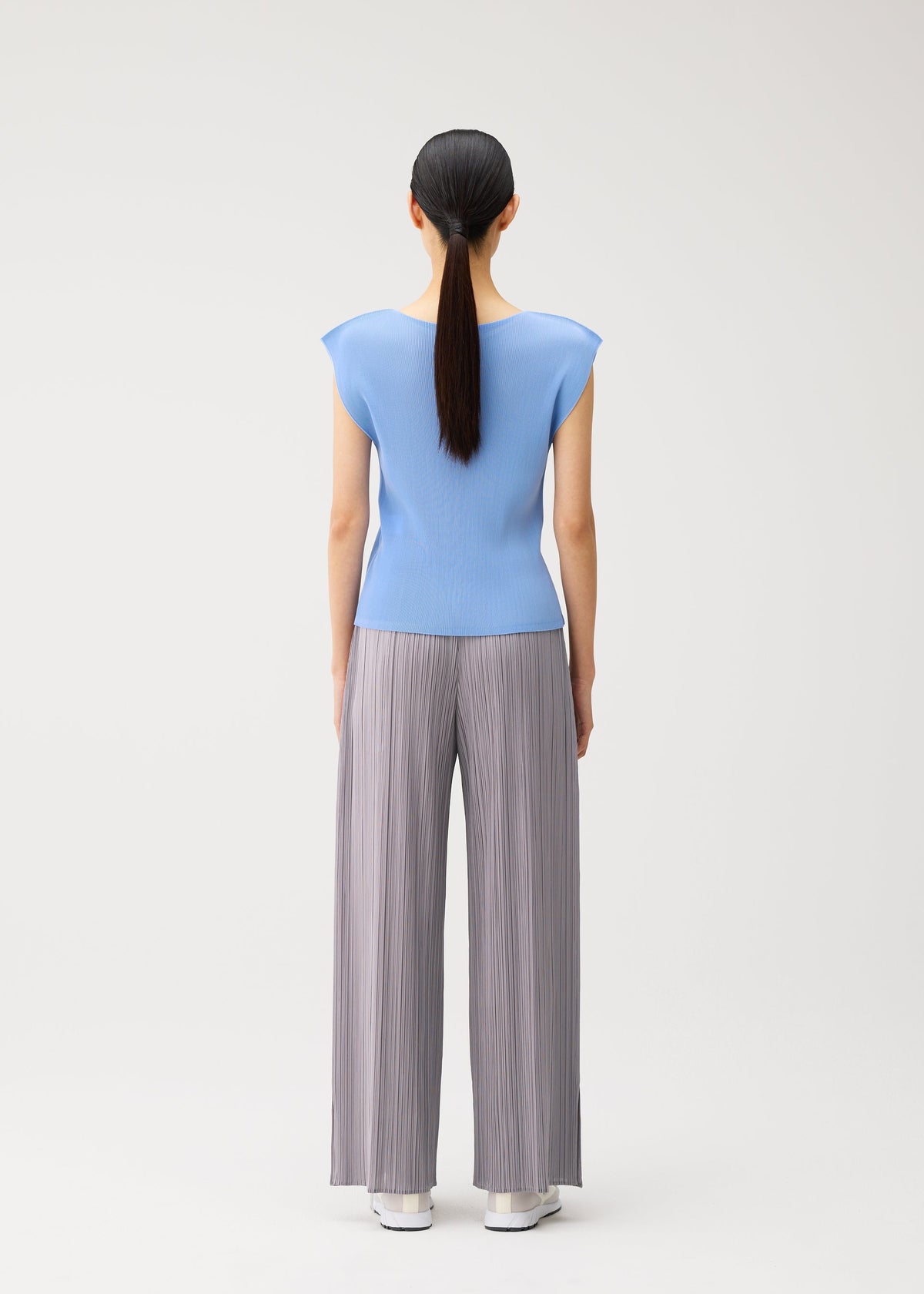 A woman stands with her back to the camera, dressed in the MIST AUGUST ensemble by PLEATS PLEASE ISSEY MIYAKE—a light blue round-neck top with French sleeves, paired with gray wide-leg pleated pants and white shoes. Her dark hair is tied back in a low ponytail against a plain white background. view 7 of 8