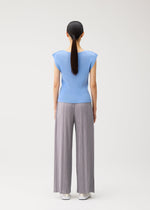 A woman stands with her back to the camera, dressed in the MIST AUGUST ensemble by PLEATS PLEASE ISSEY MIYAKE—a light blue round-neck top with French sleeves, paired with gray wide-leg pleated pants and white shoes. Her dark hair is tied back in a low ponytail against a plain white background.