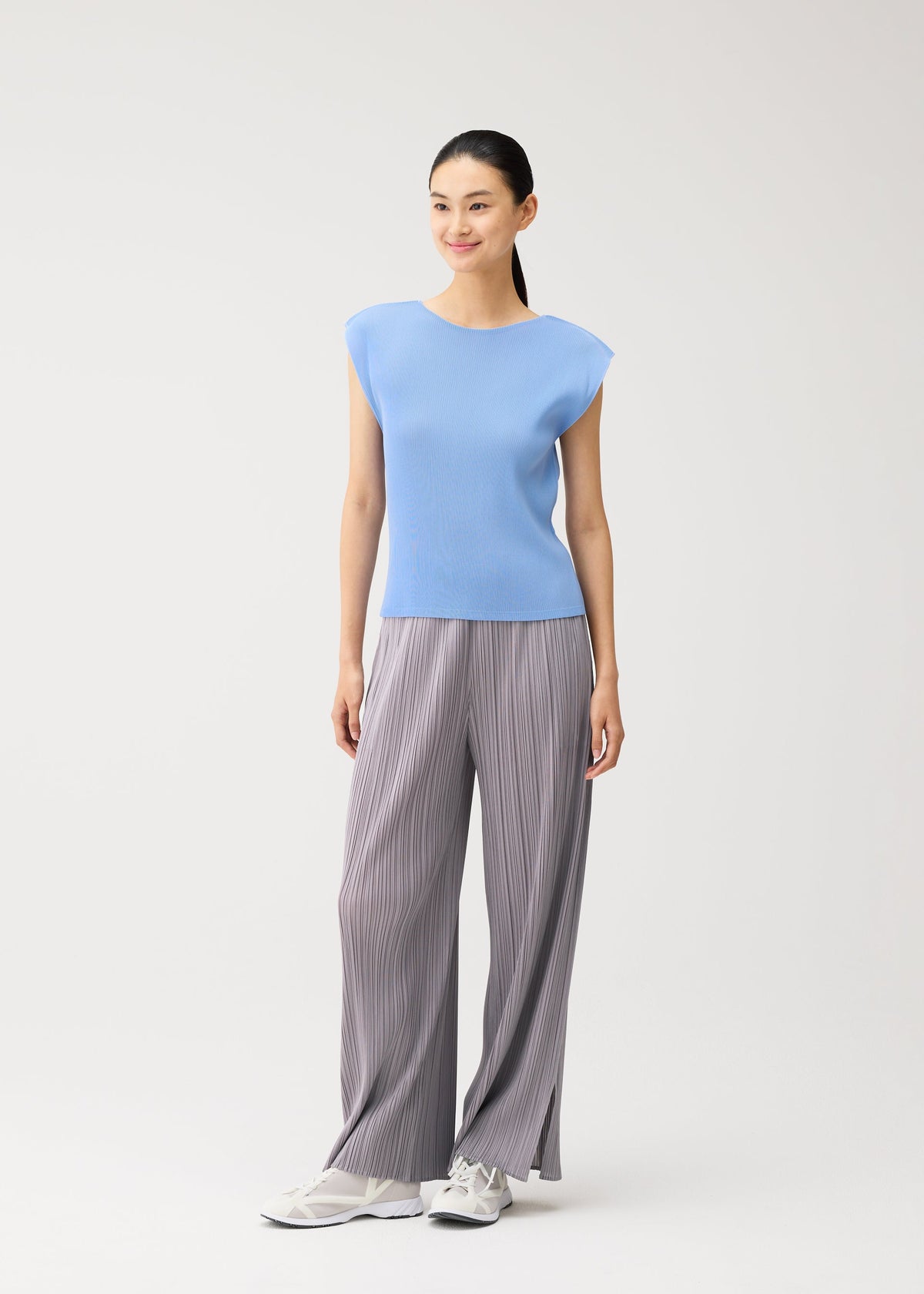 A woman stands against a plain background, smiling and facing slightly to the side, dressed in the MIST AUGUST light blue round-neck top with French sleeves and loose gray pleated pants from PLEATS PLEASE ISSEY MIYAKE, complemented by white sneakers. view 8 of 8