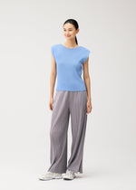 A woman stands against a plain background, smiling and facing slightly to the side, dressed in the MIST AUGUST light blue round-neck top with French sleeves and loose gray pleated pants from PLEATS PLEASE ISSEY MIYAKE, complemented by white sneakers.