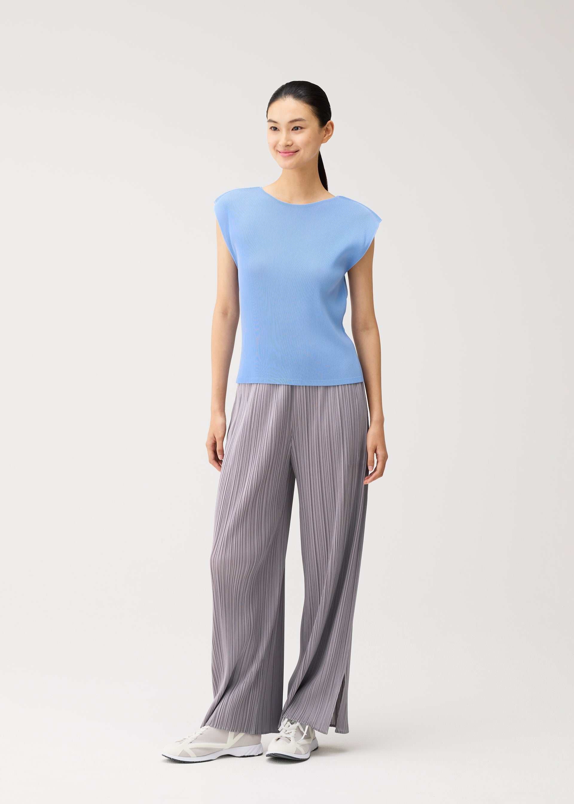 Zoomed view A woman stands against a plain background, smiling and facing slightly to the side, dressed in the MIST AUGUST light blue round-neck top with French sleeves and loose gray pleated pants from PLEATS PLEASE ISSEY MIYAKE, complemented by white sneakers.