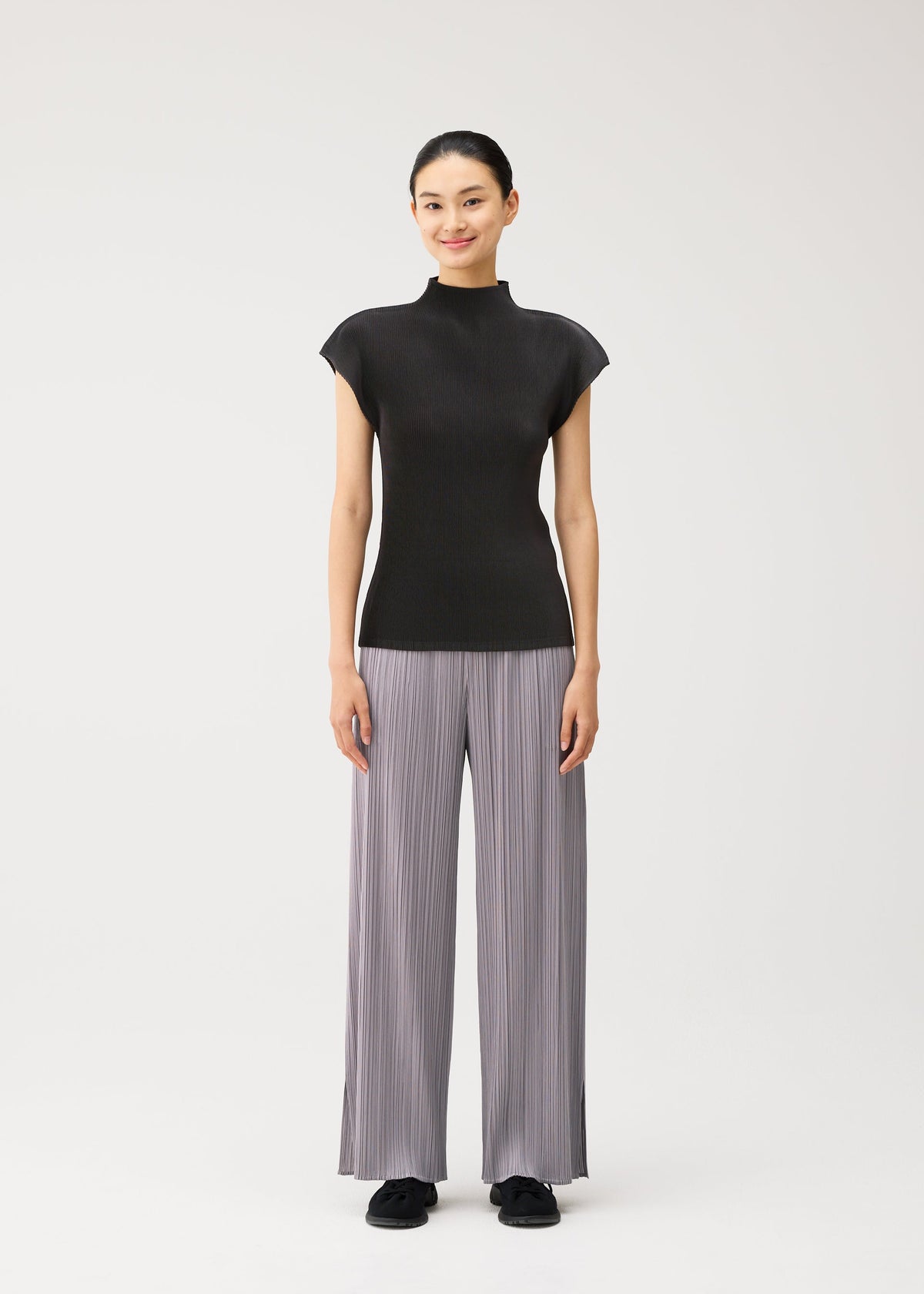 A person stands facing forward, smiling, wearing the MIST AUGUST lightweight short-sleeved black top and wide-legged light gray pleated pants from PLEATS PLEASE ISSEY MIYAKE with black shoes, against a plain white background. view 5 of 8