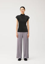 A person stands facing forward, smiling, wearing the MIST AUGUST lightweight short-sleeved black top and wide-legged light gray pleated pants from PLEATS PLEASE ISSEY MIYAKE with black shoes, against a plain white background.