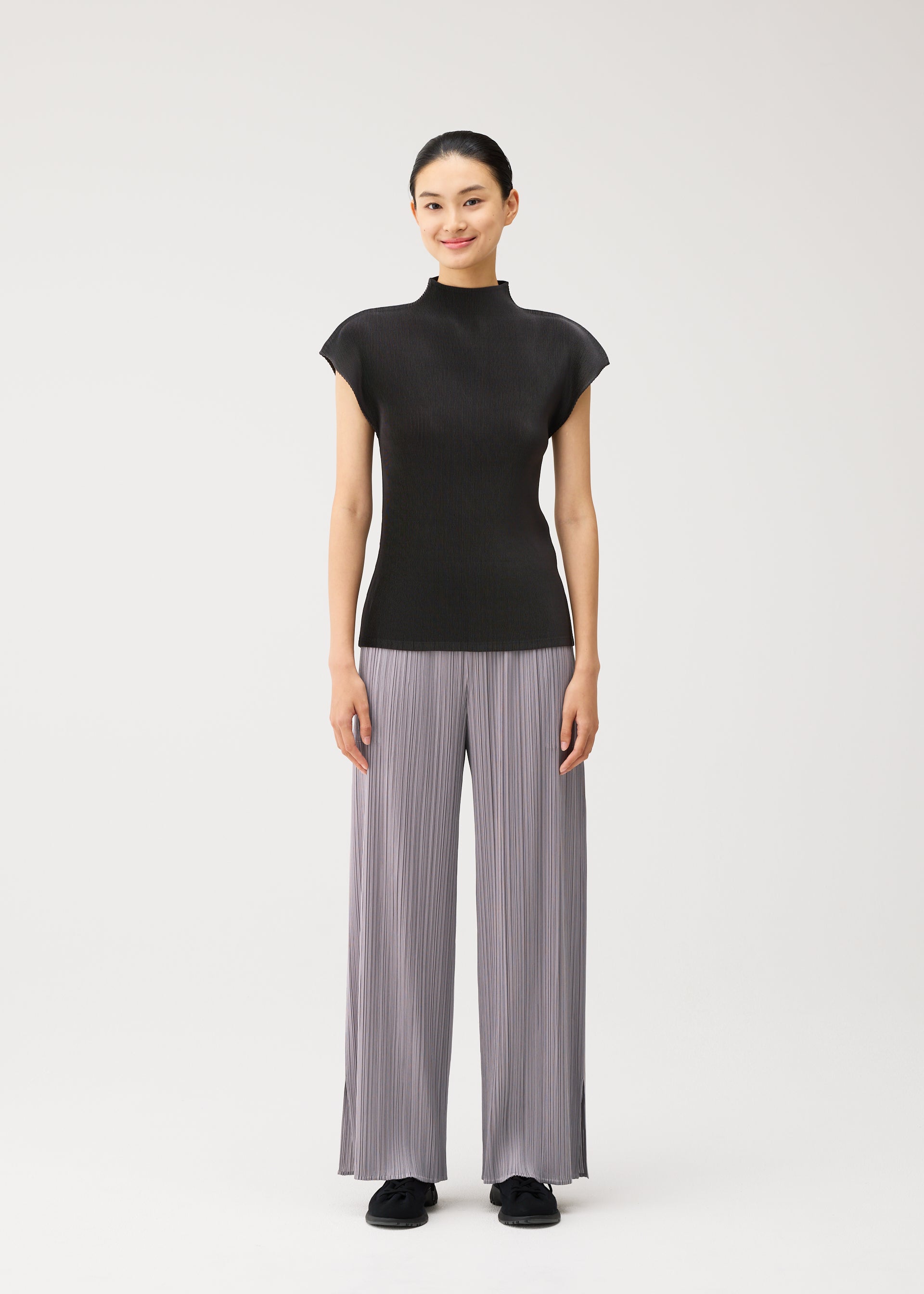 Zoomed view A person stands facing forward, smiling, wearing the MIST AUGUST lightweight short-sleeved black top and wide-legged light gray pleated pants from PLEATS PLEASE ISSEY MIYAKE with black shoes, against a plain white background.
