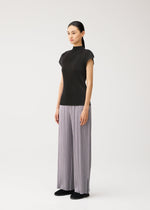 A woman stands against a plain white background wearing the MIST AUGUST lightweight black short-sleeve top and wide-leg, light gray pleated pants from PLEATS PLEASE ISSEY MIYAKE, paired with black shoes.