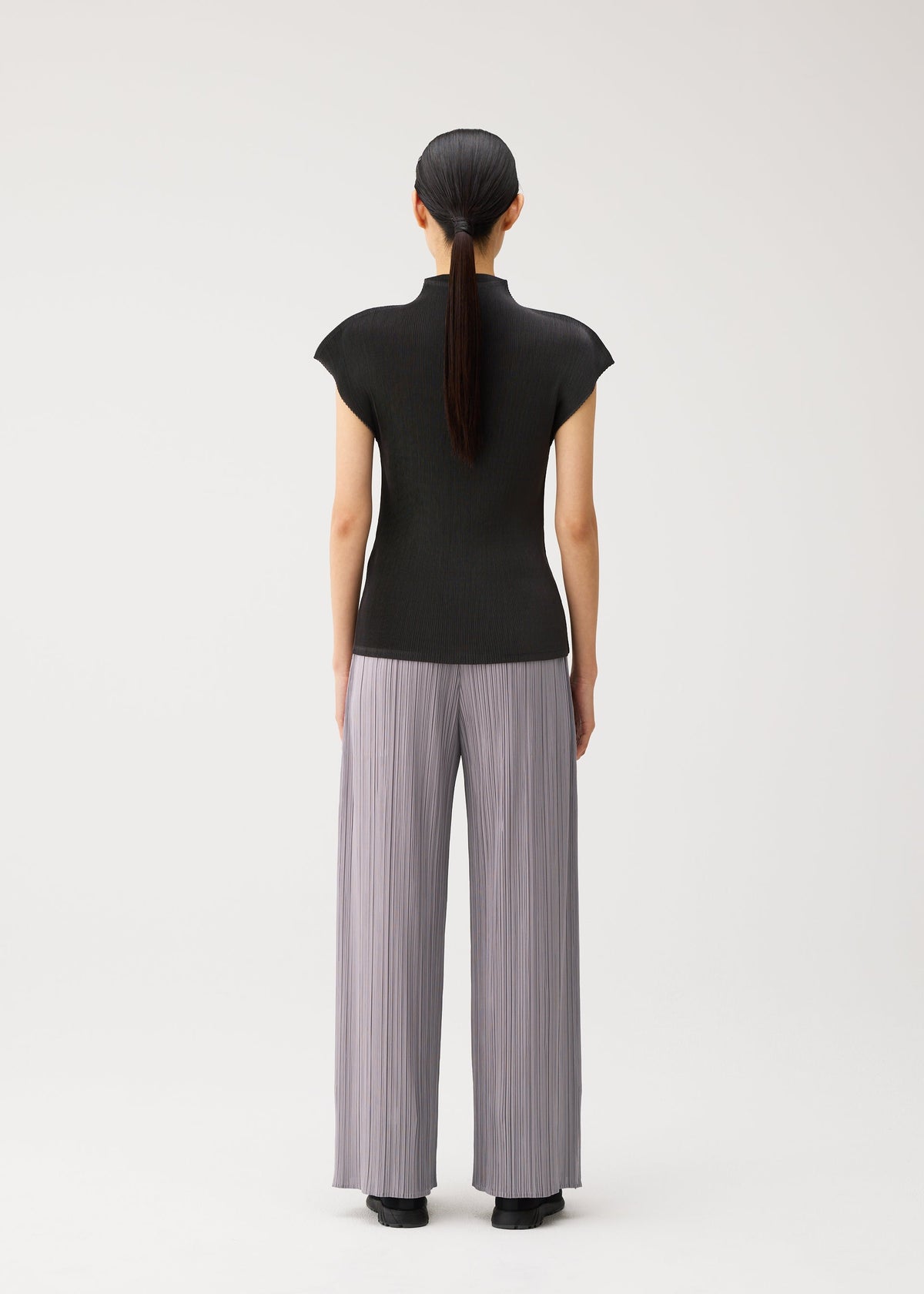 A person with long dark hair in a ponytail stands facing away, wearing the MIST AUGUST black lightweight short-sleeve top and wide, light gray pleated pants by PLEATS PLEASE ISSEY MIYAKE against a plain white background. view 7 of 8