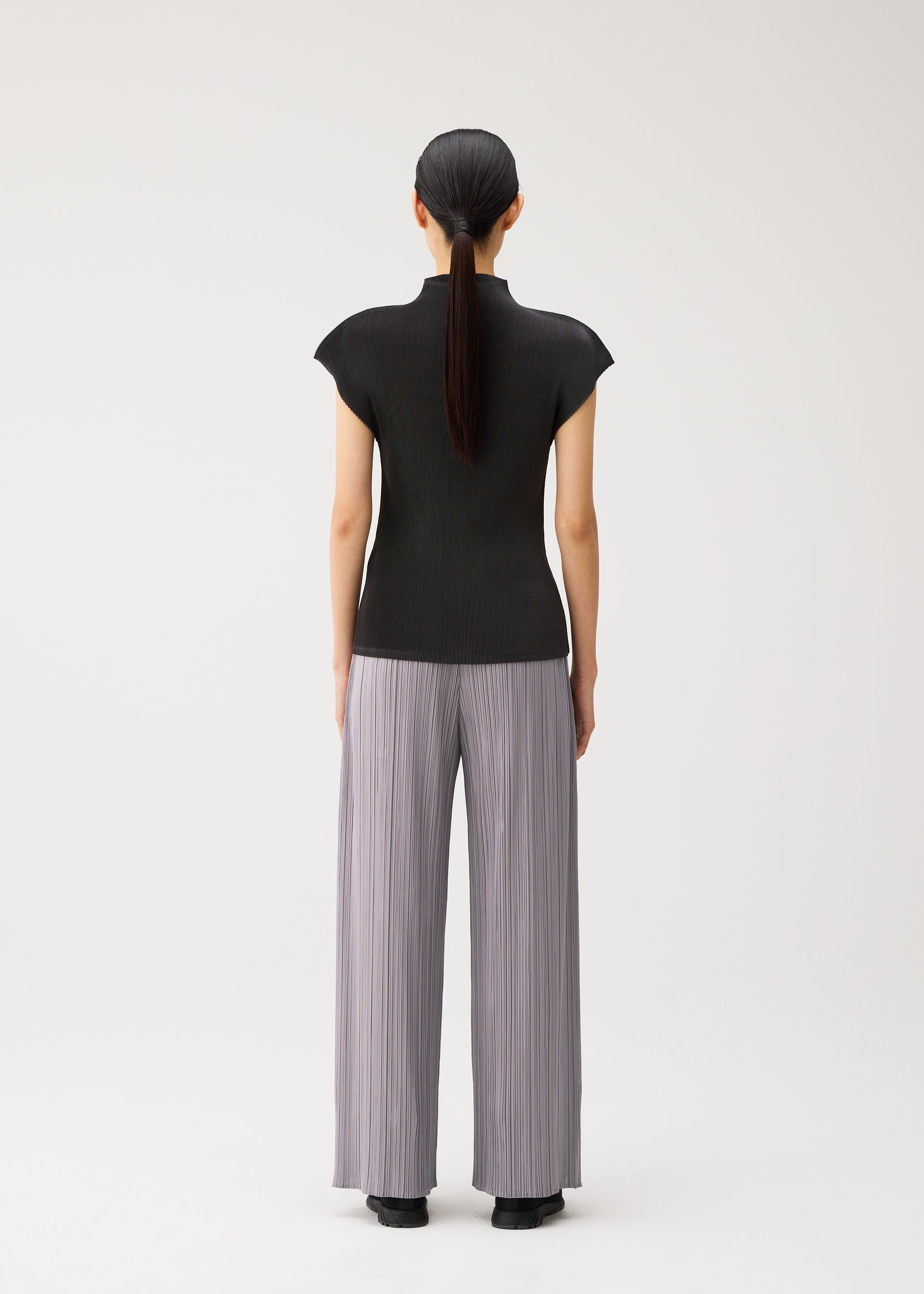 Zoomed view A person with long dark hair in a ponytail stands facing away, wearing the MIST AUGUST black lightweight short-sleeve top and wide, light gray pleated pants by PLEATS PLEASE ISSEY MIYAKE against a plain white background.