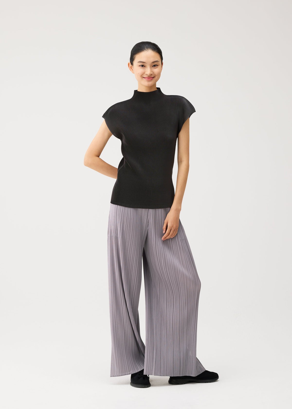 A woman stands smiling against a plain white background, wearing the MIST AUGUST lightweight black sleeveless top and wide-leg, light gray pleated pants from PLEATS PLEASE ISSEY MIYAKE, paired with black shoes. view 8 of 8
