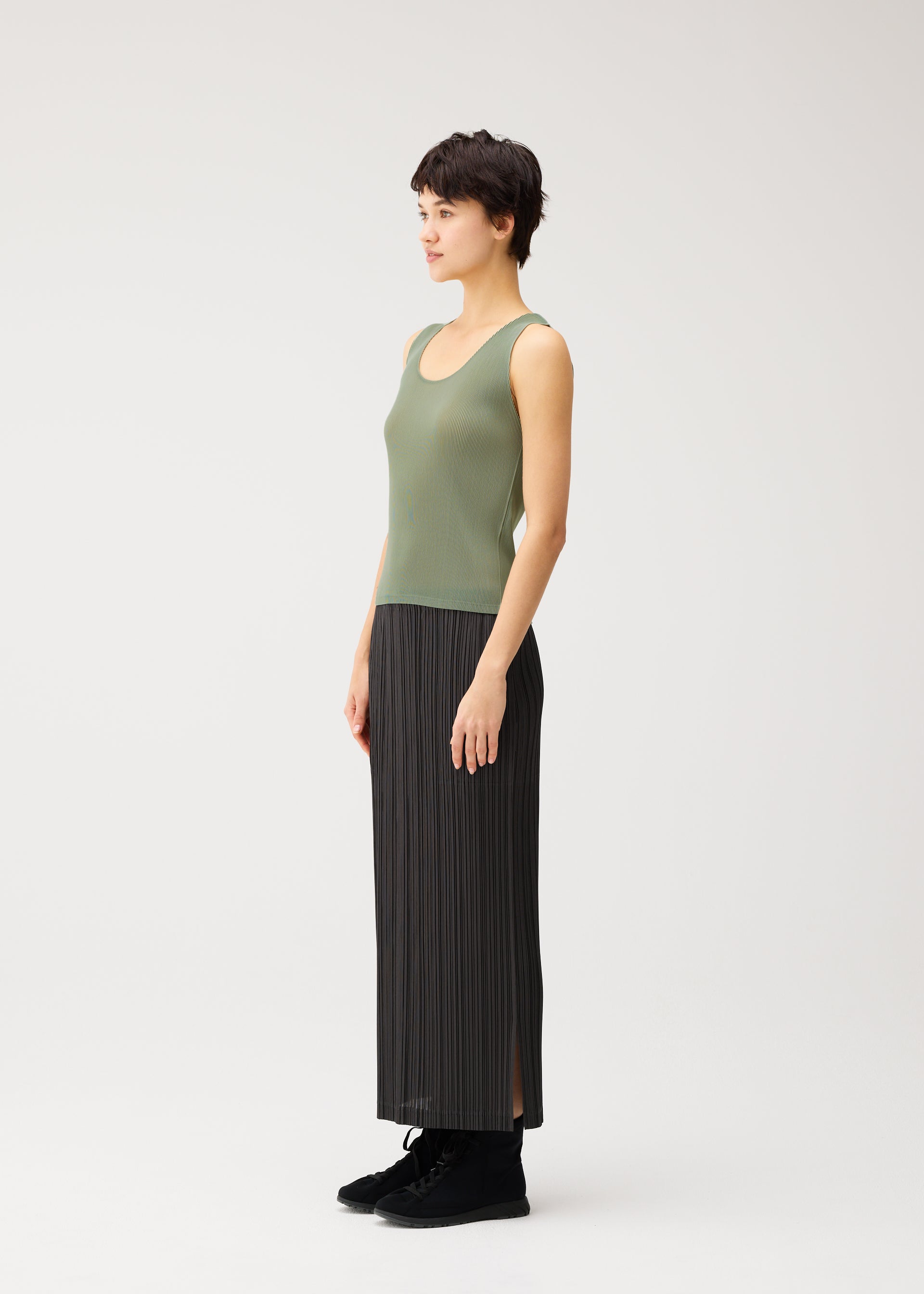 Zoomed view A woman with short dark hair stands in profile against a plain white background, wearing the MIST SEPTEMBER tank top by PLEATS PLEASE ISSEY MIYAKE—a lightweight green round-neck style—paired with a long black pleated skirt.