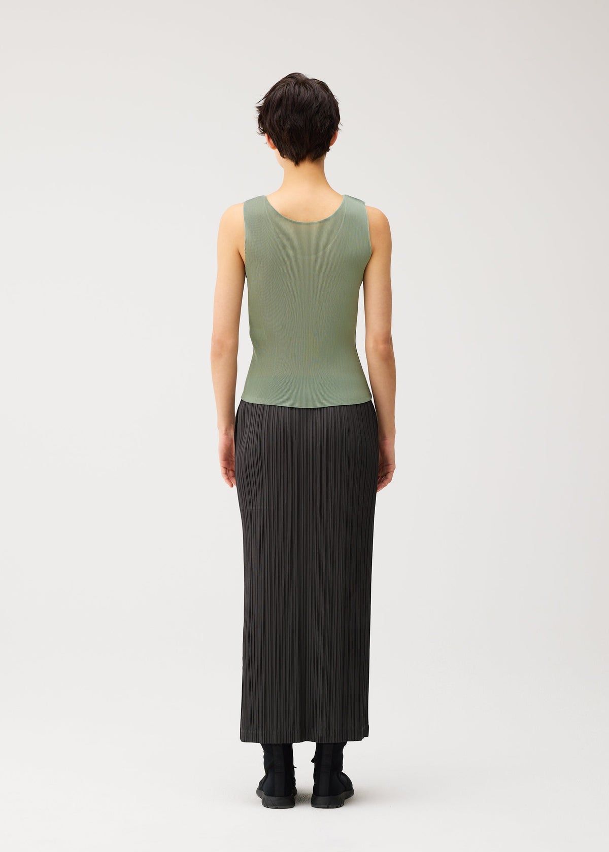 A person with short dark hair, seen from behind, is wearing the MIST SEPTEMBER lightweight green round-neck tank top and a long black pleated skirt by PLEATS PLEASE ISSEY MIYAKE, paired with black shoes, against a plain white background. view 6 of 7