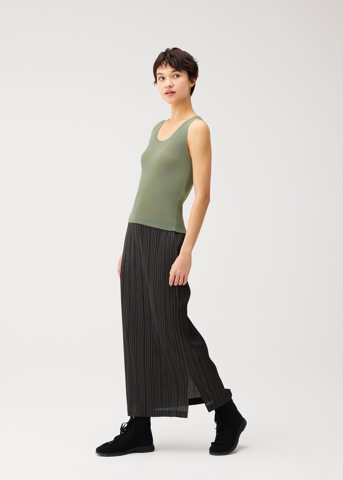 A person is standing sideways against a plain white background, wearing the MIST SEPTEMBER sleeveless green round-neck tank top and a long black pleated skirt by PLEATS PLEASE ISSEY MIYAKE, paired with black boots. view 7 of 7