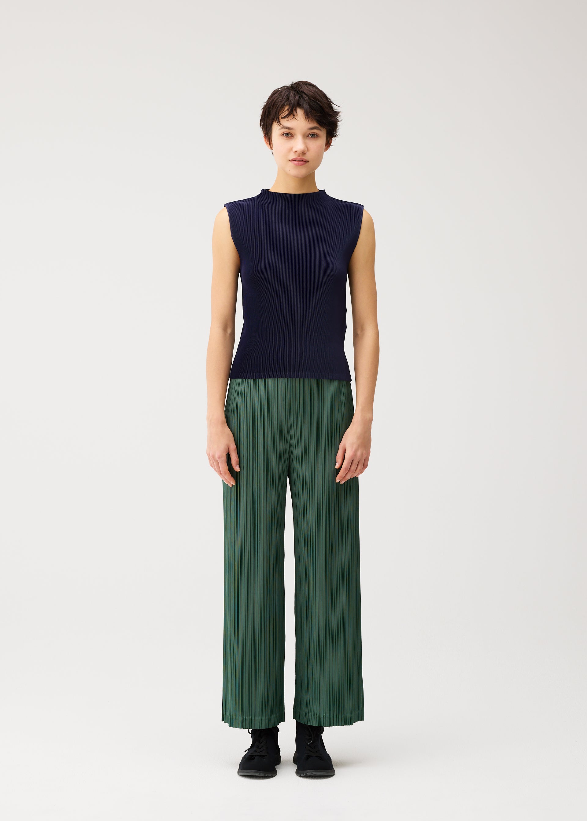 Zoomed view A person stands against a plain background, dressed in the PLEATS PLEASE ISSEY MIYAKE MIST SEPTEMBER high-neck sleeveless dark blue top and green wide-leg pleated pants with fine pleats, paired with black shoes.
