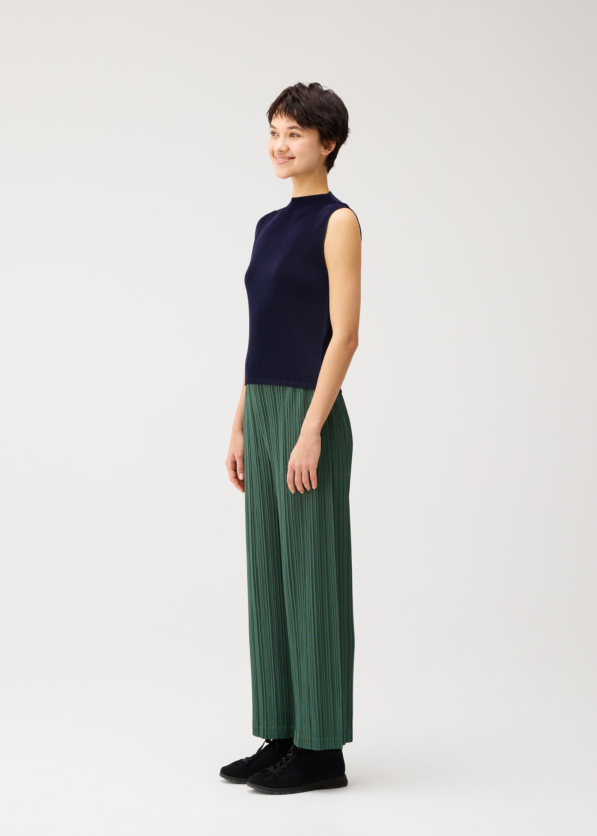 Zoomed view A woman stands sideways, smiling, wearing the MIST SEPTEMBER high-neck sleeveless navy top and loose green finely pleated pants from PLEATS PLEASE ISSEY MIYAKE, paired with black shoes against a plain light background.