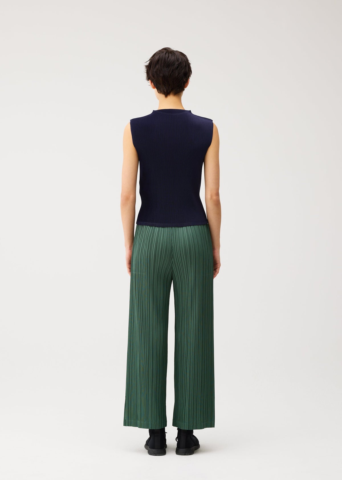 A person with short dark hair is seen from behind, wearing the MIST SEPTEMBER lightweight high-neck sleeveless navy top and loose green finely pleated pants by PLEATS PLEASE ISSEY MIYAKE, standing against a plain white background. view 6 of 7