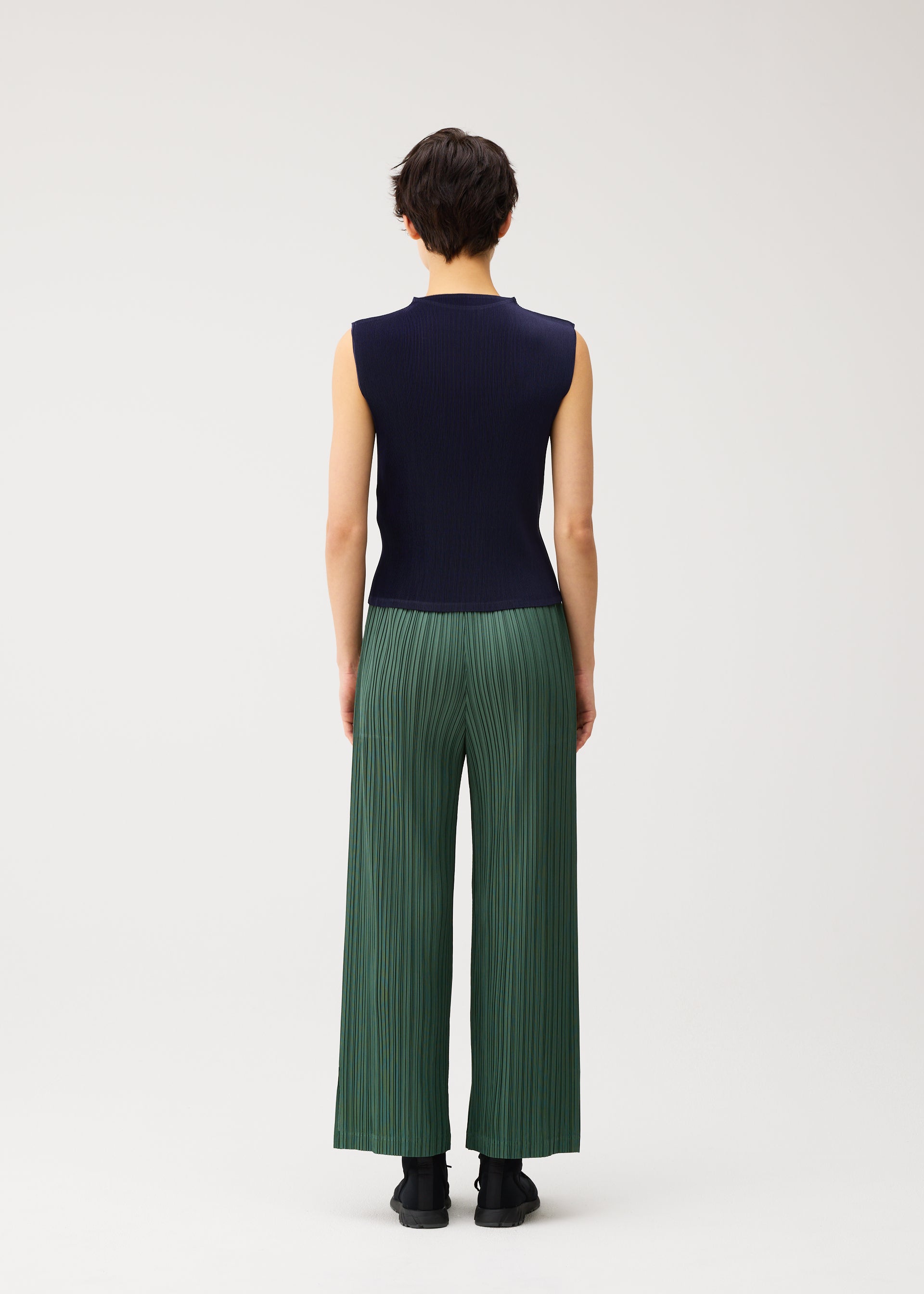Zoomed view A person with short dark hair is seen from behind, wearing the MIST SEPTEMBER lightweight high-neck sleeveless navy top and loose green finely pleated pants by PLEATS PLEASE ISSEY MIYAKE, standing against a plain white background.