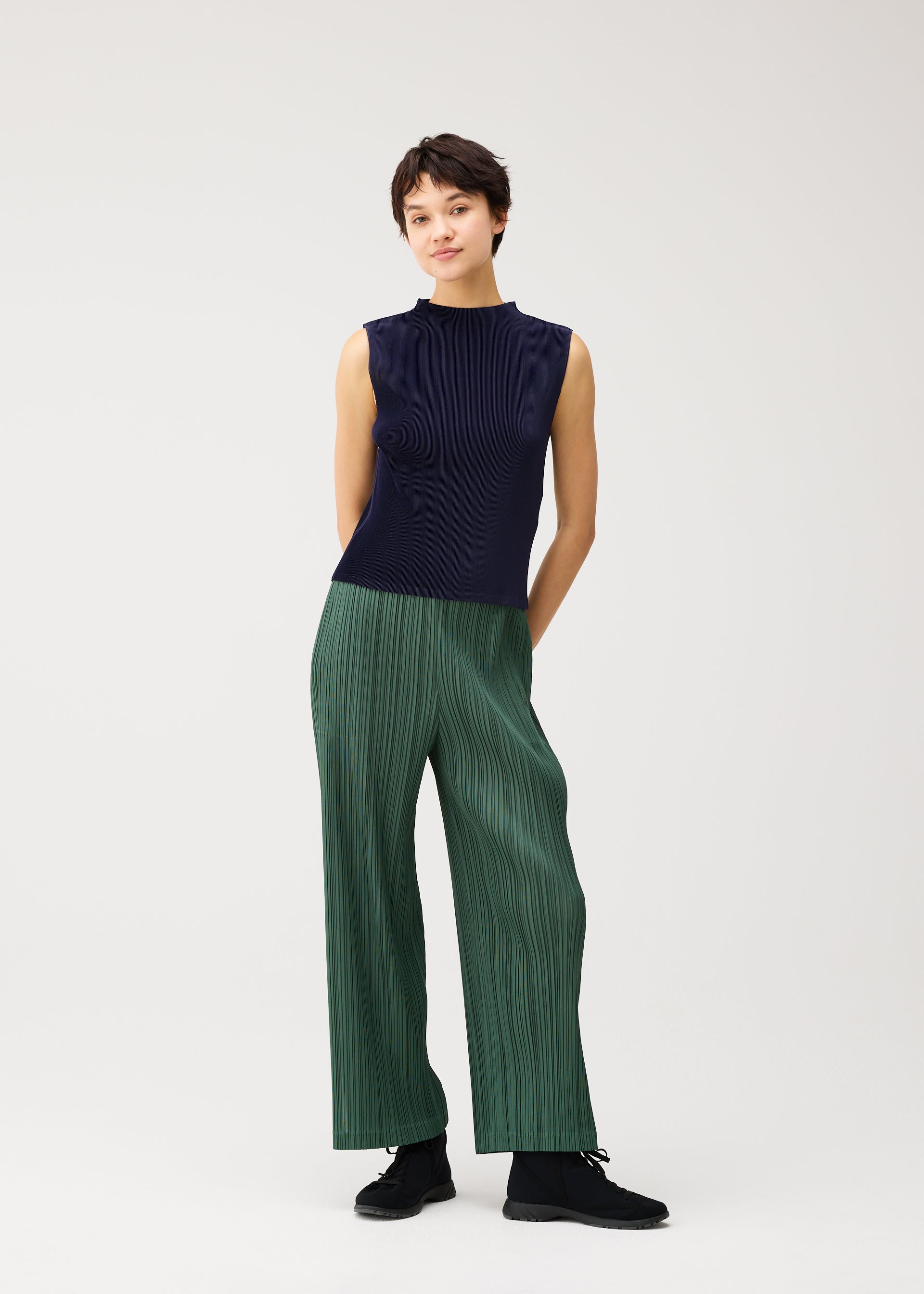 Zoomed view A person with short brown hair stands against a plain background, wearing the MIST SEPTEMBER high-neck sleeveless navy top and green wide-leg finely pleated pants from PLEATS PLEASE ISSEY MIYAKE, paired with black shoes. Their hands are positioned behind their back.