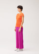 A person stands in profile against a plain background, wearing the MIST SEPTEMBER orange round-neck top and bright pink pleated pants by PLEATS PLEASE ISSEY MIYAKE, paired with white shoes.