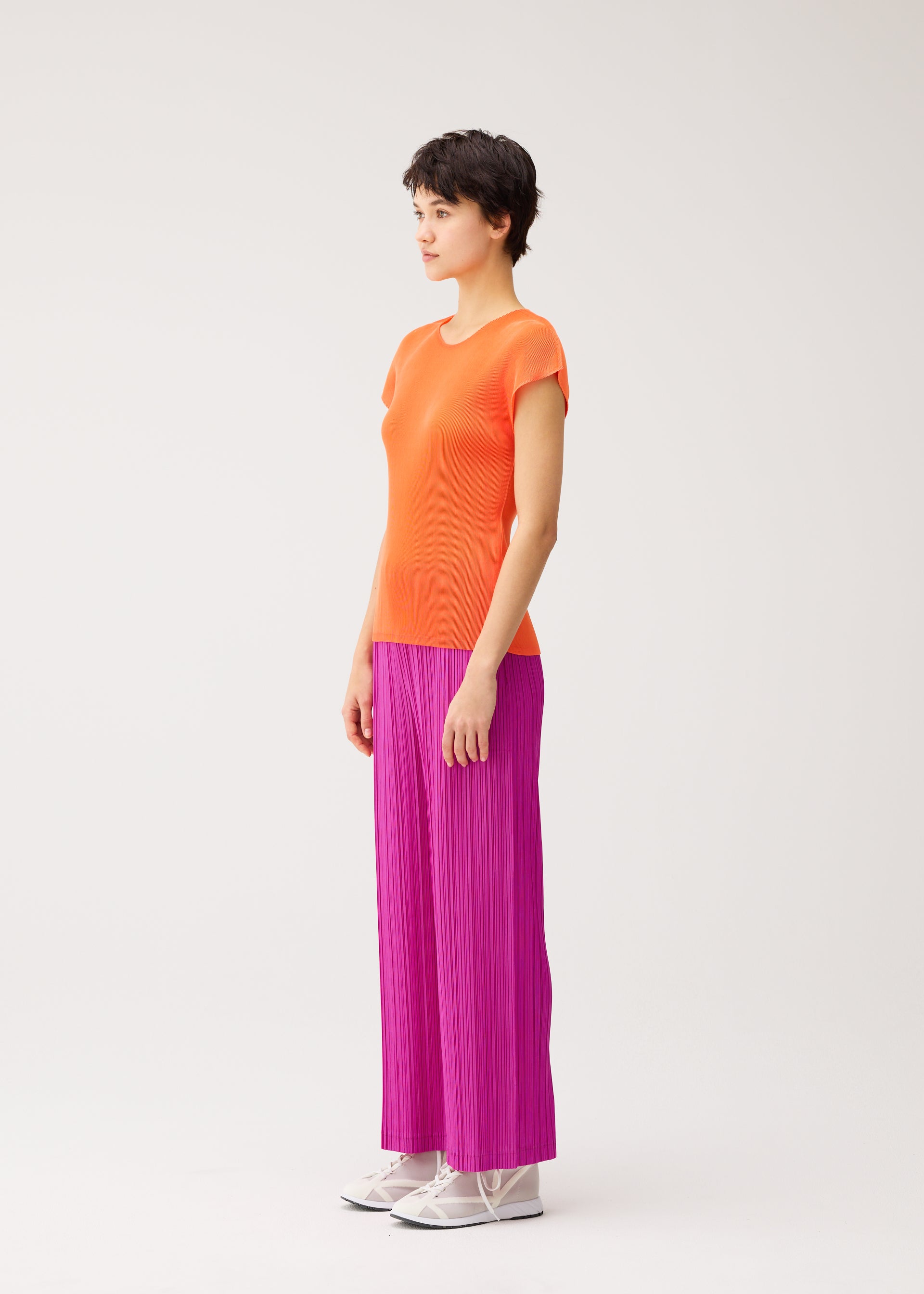 Zoomed view A person stands in profile against a plain background, wearing the MIST SEPTEMBER orange round-neck top and bright pink pleated pants by PLEATS PLEASE ISSEY MIYAKE, paired with white shoes.
