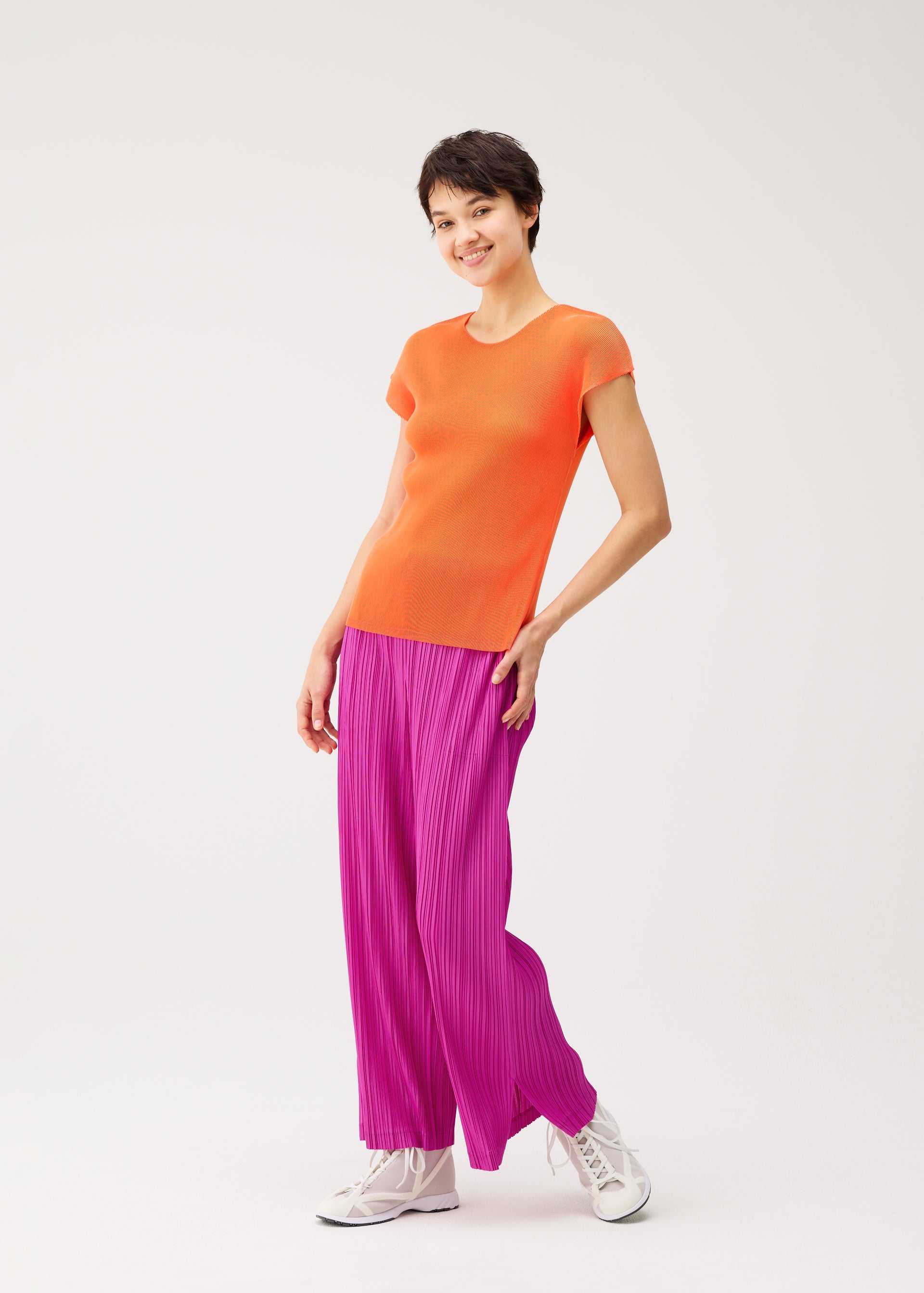 Zoomed view A person wearing the MIST SEPTEMBER top by PLEATS PLEASE ISSEY MIYAKE, styled with loose bright pink pleated pants and white sneakers, stands smiling against a plain white background.