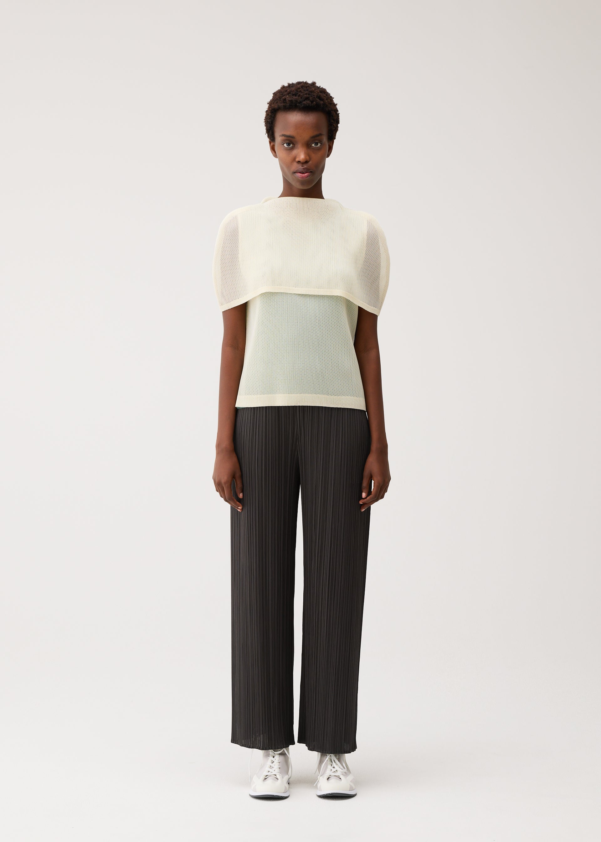 Zoomed view A person stands against a plain background, wearing the TATAMI AUGUST by PLEATS PLEASE ISSEY MIYAKE: a sheer, pale yellow boat-neck mesh top layered over a light shirt, paired with black pleated wide-leg pants and white sneakers. They have short hair and a neutral expression.