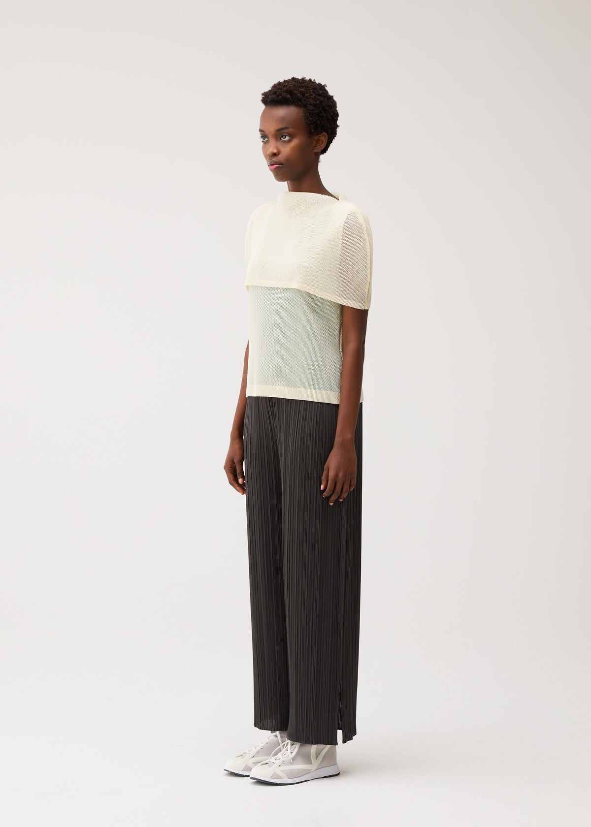 A person stands sideways against a plain light background, dressed in the PLEATS PLEASE ISSEY MIYAKE TATAMI AUGUST black wide-leg pleated pants, styled with a sheer, light yellow boat-neck top layered over a white shirt and paired with white sneakers. view 7 of 9