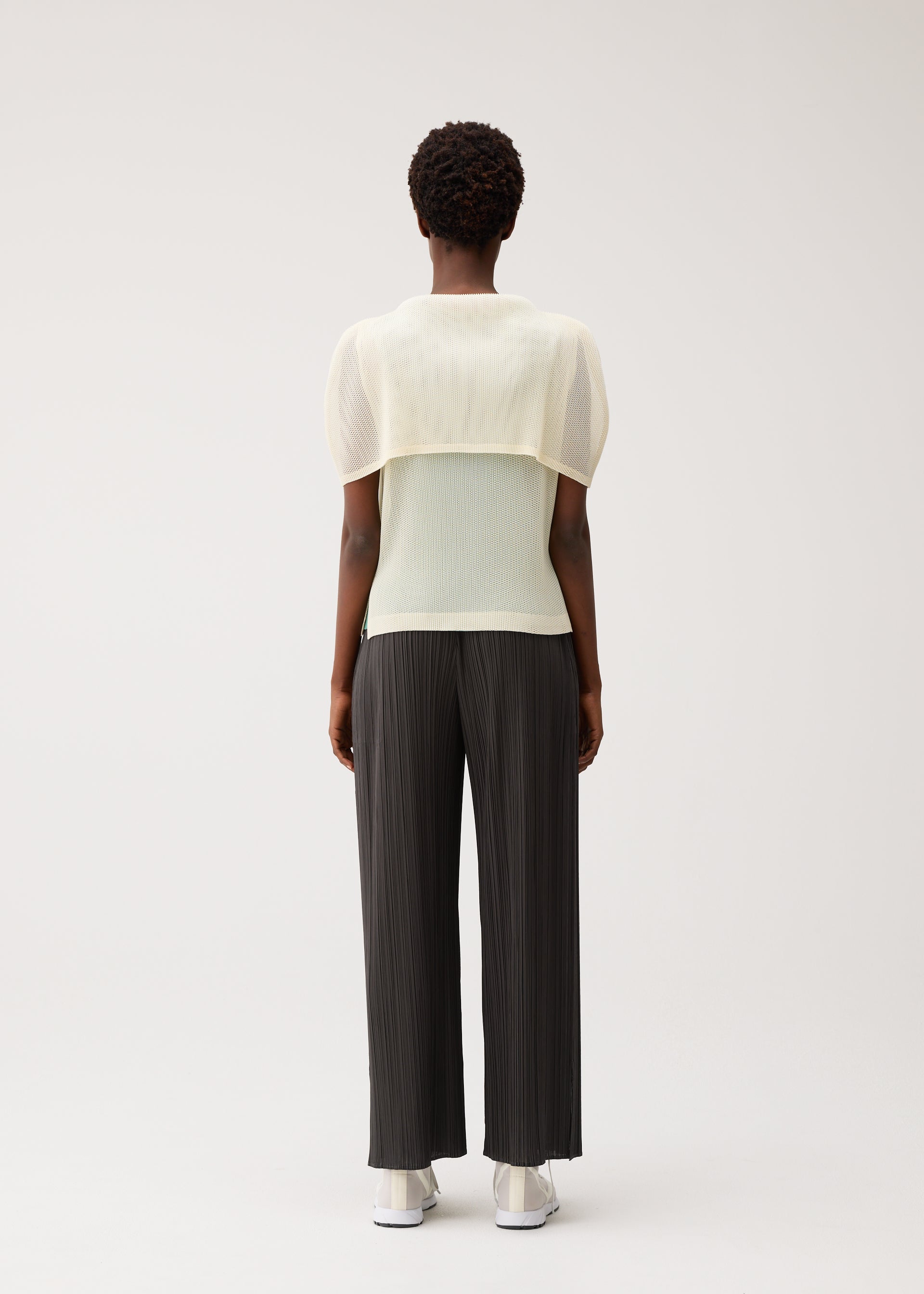 Zoomed view A person stands facing away from the camera, dressed in the TATAMI AUGUST wide-leg black pleated pants by PLEATS PLEASE ISSEY MIYAKE, paired with a sheer, short-sleeve cream mesh top and white shoes against a plain light background.