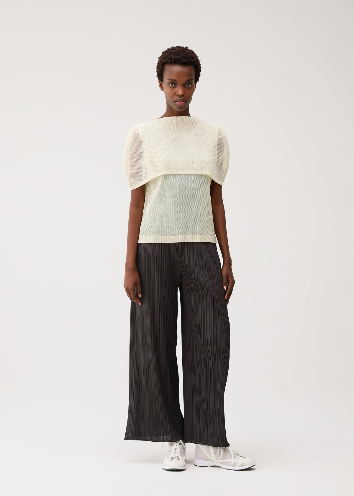 A person stands against a plain light background wearing the TATAMI AUGUST black pleated wide-leg pants by PLEATS PLEASE ISSEY MIYAKE, paired with a sheer, light-colored mesh fabric top with wide, structured sleeves and white sneakers. view 9 of 9