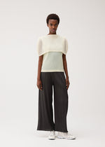 A person stands against a plain light background wearing the TATAMI AUGUST black pleated wide-leg pants by PLEATS PLEASE ISSEY MIYAKE, paired with a sheer, light-colored mesh fabric top with wide, structured sleeves and white sneakers.