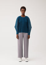A person stands against a plain background, facing forward with a neutral expression, wearing the TATAMI AUGUST button-up top in teal—one of several cool-feeling colors—from PLEATS PLEASE ISSEY MIYAKE. The top features sheer sleeves and is paired with loose light gray pleated pants and white shoes.