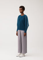 A person wearing the TATAMI AUGUST cool-feeling teal button-up cardigan and light gray pleated wide-leg pants from PLEATS PLEASE ISSEY MIYAKE, paired with white shoes, stands against a plain light background, looking slightly to the left.