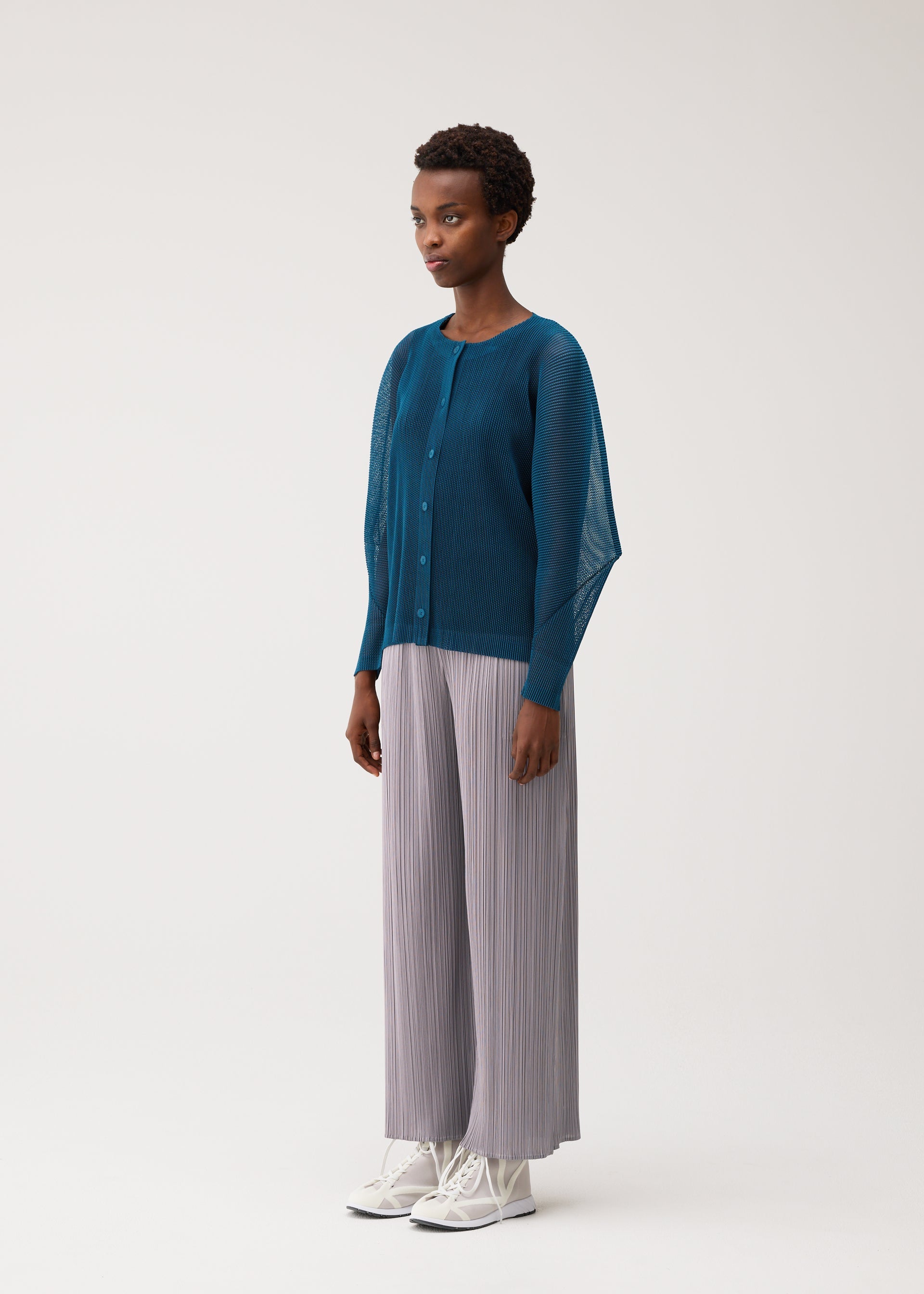 Zoomed view A person wearing the TATAMI AUGUST cool-feeling teal button-up cardigan and light gray pleated wide-leg pants from PLEATS PLEASE ISSEY MIYAKE, paired with white shoes, stands against a plain light background, looking slightly to the left.