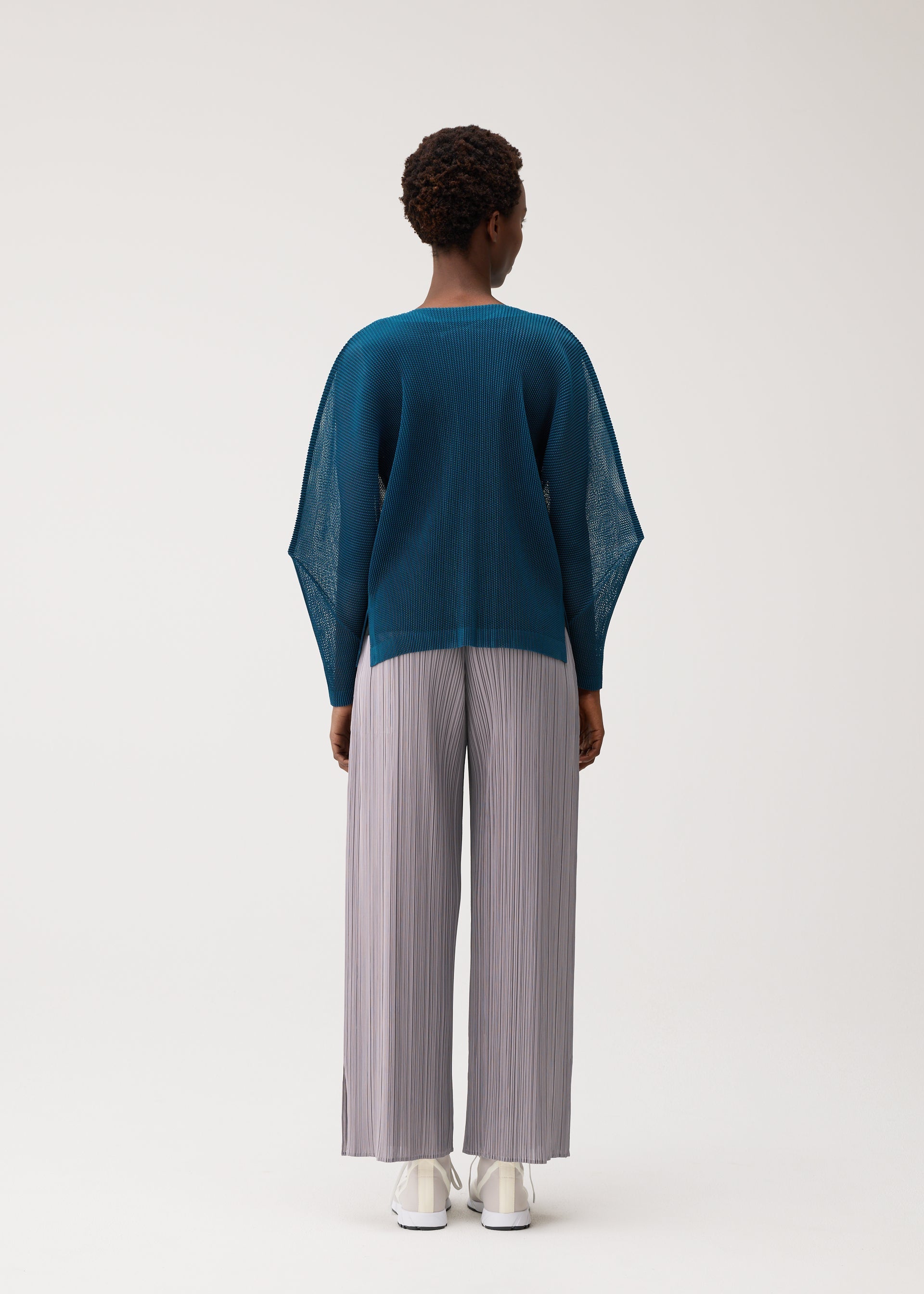 Zoomed view Wearing the TATAMI AUGUST wide-leg, light gray pleated pants from PLEATS PLEASE ISSEY MIYAKE, a person stands facing away in a loose teal sweater and crisp white shoes against a plain white background, effortlessly pairing cool tones and airy textures.