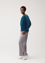 A woman stands in profile wearing a teal sheer long-sleeve top layered beneath a pleated mesh cardigan, light gray pleated wide-leg pants, and white sneakers against a plain light background. The ensemble showcases cool-feeling colors for an effortlessly modern look.