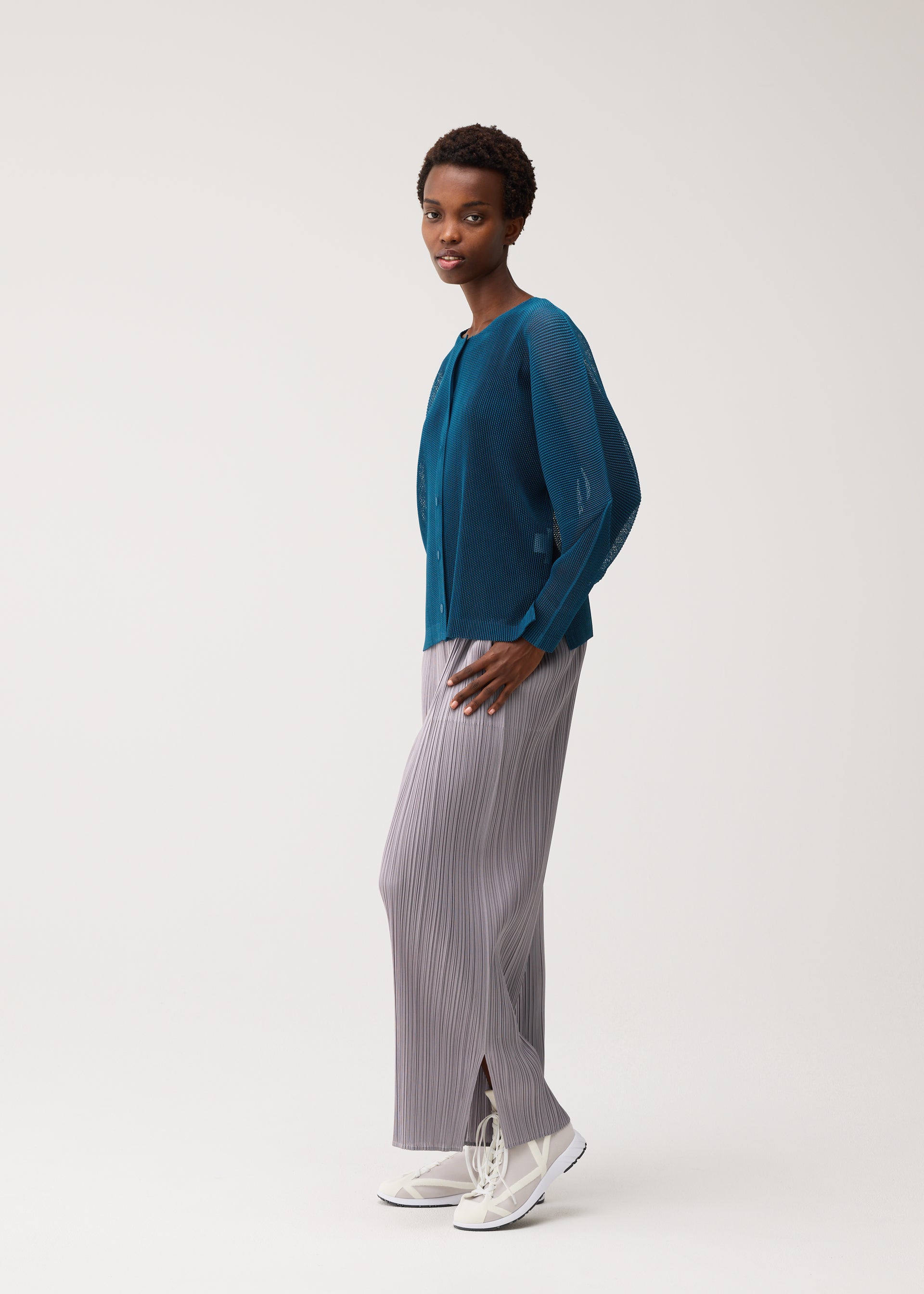 Zoomed view A woman stands in profile wearing a teal sheer long-sleeve top layered beneath a pleated mesh cardigan, light gray pleated wide-leg pants, and white sneakers against a plain light background. The ensemble showcases cool-feeling colors for an effortlessly modern look.