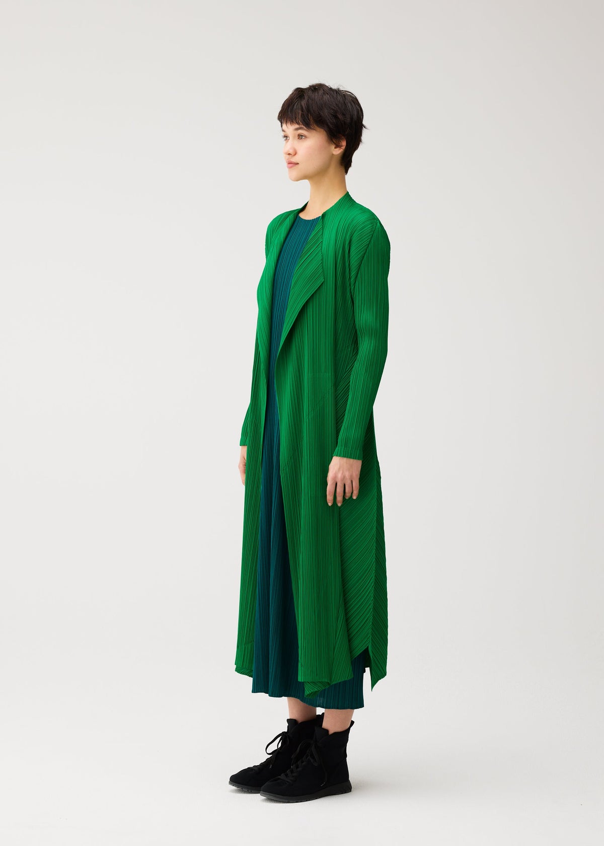 A person with short dark hair stands in profile, wearing the MONTHLY COLORS : OCTOBER coat by PLEATS PLEASE ISSEY MIYAKE over a blue dress and black boots against a plain white background. view 9 of 11